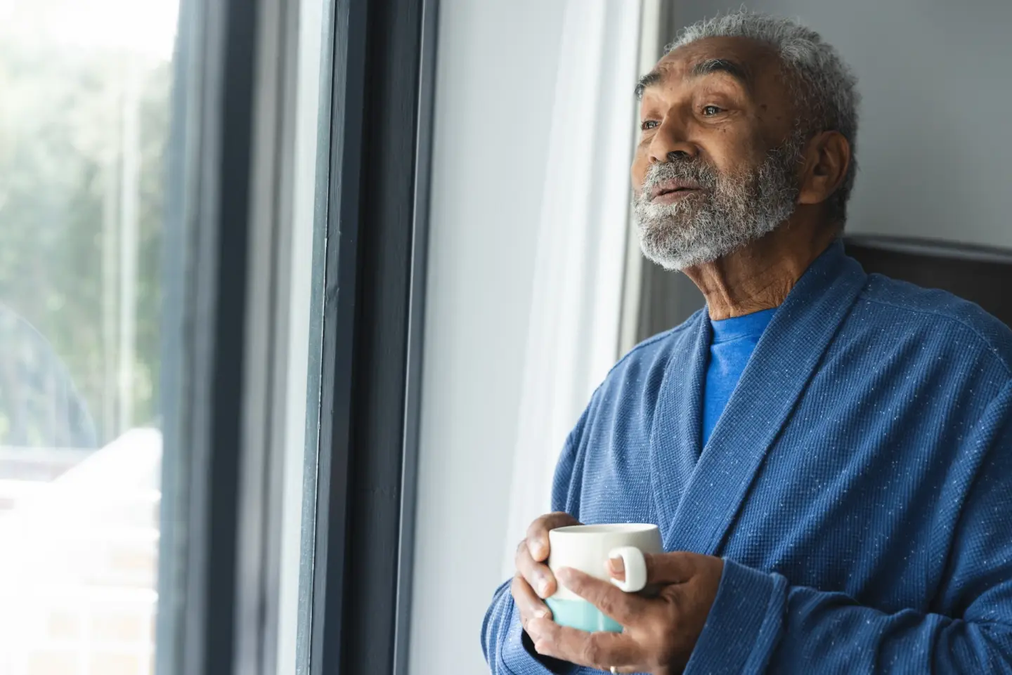 thoughtful senior biracial man wearing bathrobe holding mug coffee by window home